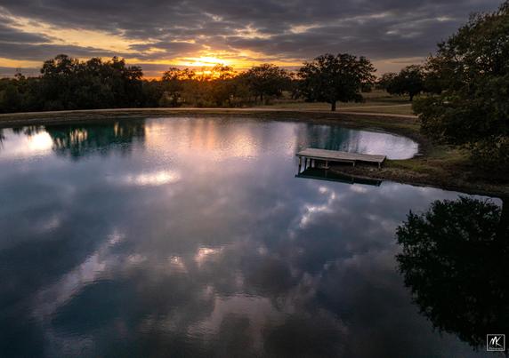 Color photo of the sun setting near the horizon in a partly cloudy sky. In the foreground is a large pond with the sky reflected in the water.