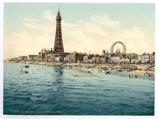 The image depicts a vintage postcard featuring an idyllic coastal scene from Central Pier in Blackpool, England. The prominent structures include the tall Victorian-era tower known as The Tower and a Ferris wheel with gondola-style carriages on its circular structure, suggesting it was taken during that era's amusement rides popularity.

In the foreground, calm blue waters gently lap against an expansive sandy beach where people are scattered about; some near the waterline while others stroll along. A sailboat is anchored close to shore, and a small pier extends into the sea with more individuals on or around it. The town in the background consists of various buildings indicative of late 19th-century architecture.

The overall color tone appears aged and faded, typical for such vintage postcards from that period. Text at the bottom indicates "Blackpool - From Central Pier," along with a date likely signifying when this photo was created or is intended to represent (11/38). The image captures an early 20th-century representation of Blackpool's seaside attractions and leisure activities, reflecting its status as a popular destination during that time.