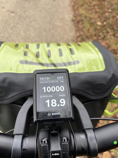 Image shows a close-up of a bicycle's handlebar with a display. The screen indicates a total distance of 10,000 km and an average speed of 18.9 km/h. The background features a blurred pathway with foliage.

Das Bild zeigt eine Nahaufnahme des Lenkers eines Fahrrads mit einem Display. Der Bildschirm zeigt eine Gesamtstrecke von 10.000 km und eine Durchschnittsgeschwindigkeit von 18,9 km/h an. Der Hintergrund zeigt einen verschwommenen Weg mit Laub.