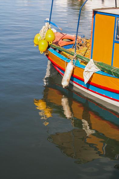 A colorful boat with yellow buoys and fishing gear is moored. The boat's reflection is visible in the still water.