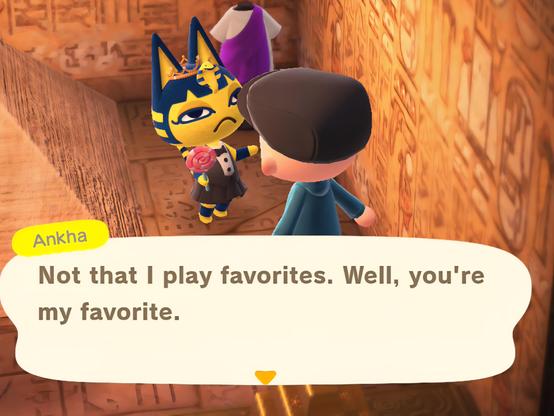 Animal Crossing: New Horizons screenshot. Image set inside of a pyramid with a smaller pyramid visible to the left. Ankha (yellow cat with blue hair and blue around her ears) wearing a golden prom crown and a tuxedo dress is talking to a bald human in a paperboy cap and blue hoodie as seen from behind. Ankha is also holding a large lollipop. In a large text box near the bottom, it shows Ankha speaking as she says, "Not to play favorites. Well, you're my favorite."