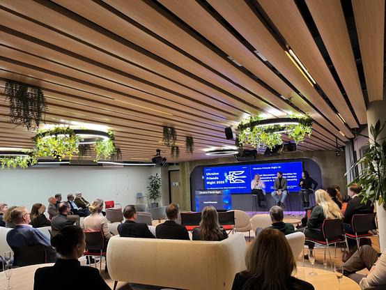 A conference or discussion event titled "Ukraine Friends Night 2023" is taking place in a modern setting. Attendees are seated, facing a stage where three speakers are engaged in discussion. The space features a wooden ceiling with hanging plants, bright lighting