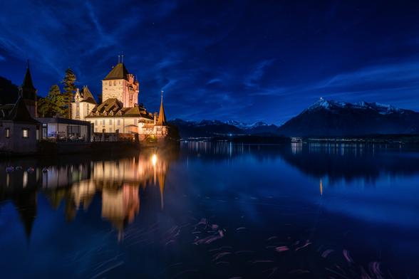 Eine Langzeitbelichtung fängt die nächtliche Szenerie des Schlosses Oberhofen am Thunersee ein. Die warmen Lichter des Schlosses bilden einen Kontrast zum tiefblauen Himmel und dem ruhigen Wasser, in dem sich die Architektur und die fernen Berglichter spiegeln. Die Aufnahme betont die zeitlose Schönheit der historischen Stätte und die beeindruckende Naturkulisse der Schweizer Alpen.