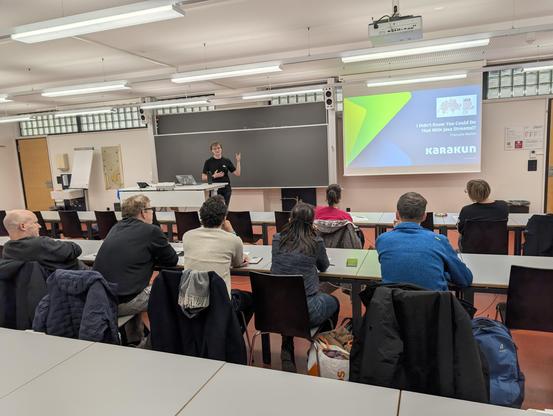 Das Foto zeigt die Teilnehmer von hinten wie sie auf den Speaker und die Leinwand mit den Slides schauen.