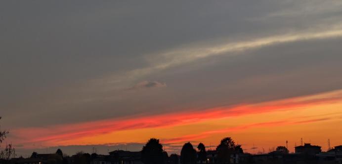 L'immagine mostra un suggestivo tramonto, caratterizzato da un forte contrasto tra i colori caldi all'orizzonte e le tonalità più scure del cielo sovrastante.
La parte inferiore del cielo è dominata da intense sfumature di rosso e arancione brillante, che si fondono gradualmente verso l'alto. 
Salendo, i colori caldi lasciano il posto a nuvole grigie e blu scuro, con una lunga striscia di nuvola bianca o grigio chiaro che attraversa diagonalmente la parte superiore del cielo.
Una silhouette scura di alberi e tetti di edifici si staglia nettamente contro i colori luminosi del tramonto, offrendo un senso di profondità e contesto urbano o semi-urbano.