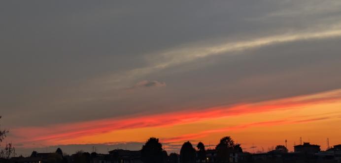 L'immagine mostra un suggestivo tramonto, caratterizzato da un forte contrasto tra i colori caldi all'orizzonte e le tonalità più scure del cielo sovrastante.
La parte inferiore del cielo è dominata da intense sfumature di rosso e arancione brillante, che si fondono gradualmente verso l'alto. 
Salendo, i colori caldi lasciano il posto a nuvole grigie e blu scuro, con una lunga striscia di nuvola bianca o grigio chiaro che attraversa diagonalmente la parte superiore del cielo.
Una silhouette scura di alberi e tetti di edifici si staglia nettamente contro i colori luminosi del tramonto, offrendo un senso di profondità e contesto urbano o semi-urbano.