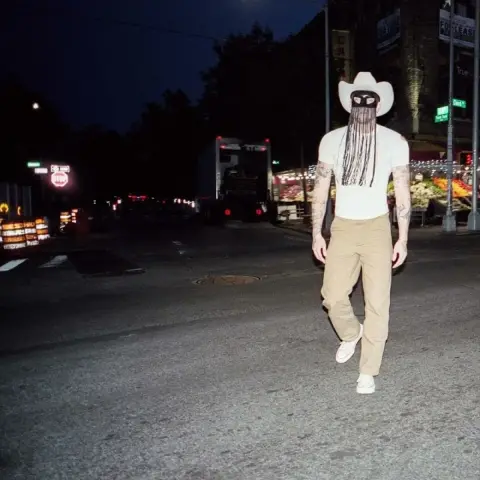 A man in a white t-shirt, khaki pants, and white sneakers walks down a pre-dawn street. He wears a black mask with fringe and a white cowboy hat, looking directly in the camera. The flash overexposes the image.