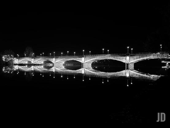 Una fotografía nocturna en blanco y negro de un puente arqueado sobre el agua. El puente presenta múltiples arcos semicirculares y está brillantemente iluminado por una fila de farolas encendidas que se extienden a lo largo de su parte superior. El agua, muy tranquila, crea un reflejo casi perfecto del puente y sus luces, lo que resulta en una composición simétrica que forma una figura similar a un símbolo de infinito o un ocho horizontal. El cielo y el agua circundante son completamente oscuros, resaltando el brillo del puente y sus reflejos.
