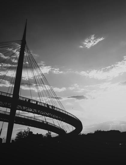 Foto orizzontale in bianco e nero del Ponte del Mare a Pescara: la struttura curva del ponte Ăš in silhouette, vista di lato, con alcune persone che camminano sul passaggio sopraelevato. Un pilone alto e i cavi del ponte salgono verso lâalto a sinistra, mentre sullo sfondo il cielo Ăš pieno di nuvole leggere e luce diffusa del tramonto. In basso si intravedono i profili scuri degli edifici e degli alberi.