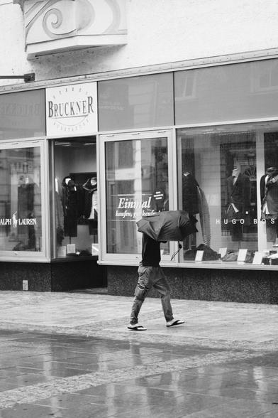 Das Foto zeigt eine Straßenszene in Schwarz-Weiß in der St. Pöltener Innenstadt an einen regnerischen windigen Tag. Man sieht eine Person mit Regenschirm der durch den Wind verbogen ist.