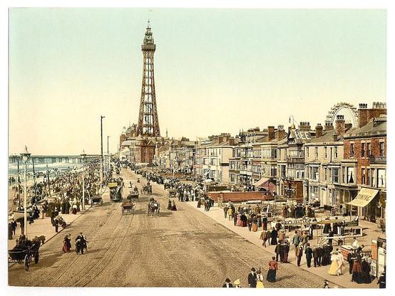 The image depicts a bustling seaside promenade in early 20th-century Britain, with the iconic tall structure resembling Blackpool Tower dominating the skyline. The street is teeming with people dressed in period attire, engaging in various activities such as walking along the promenade and socializing at outdoor booths that offer refreshments or games.

Horse-drawn carriages are interspersed among pedestrians, indicating a blend of leisurely travel methods during this era. On either side of the street, there's an array of shops with ornate facades and large windows showcasing merchandise to passersby. The architecture features steep gabled roofs common in British seaside towns.

In the background beyond the promenade is what appears to be Blackpool Pier, recognizable by its distinctive structure that juts into the sea. Other amusement structures like a Ferris wheel are also visible further down along the coast. The sky has a vintage yellowish tint typical of photochrom prints from this period, enhancing the nostalgic feel of the scene.

Overall, it captures a vibrant and leisurely atmosphere reflective of late Victorian or Edwardian England's seaside resorts' allure for tourists seeking recreation by the sea.