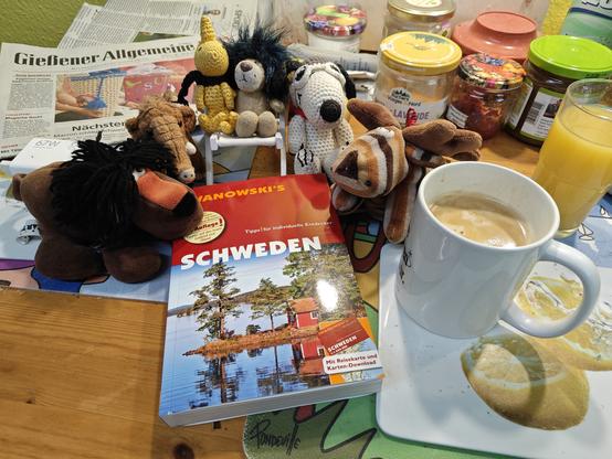 Verschiedene Plüschtiere haben sich um einen Reiseführer für Schweden versammelt, der auf dem Frühstückstisch liegt.