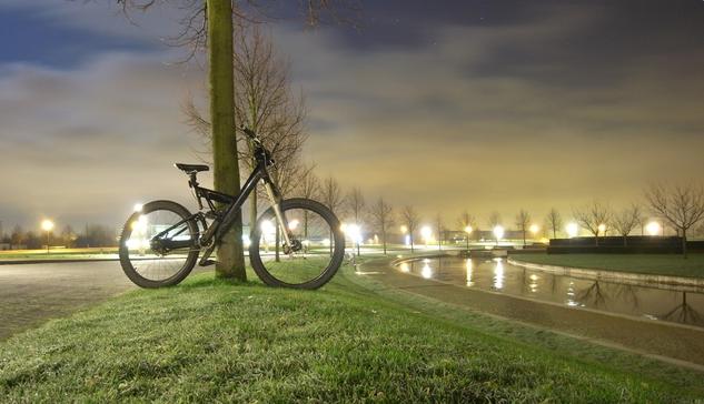 A mountain bike with a silver Lyric 16 cm suspension fork, an all-black Cannondale SuperV frame without a logo, a black Rohloff hub, and a handlebar-adjustable seatpost—not standard in 2007—and a 1400-lumen camping lamp with a tent pole mount (which couldn't be sold as a bicycle light at the time) leans against a bare tree on a frost-covered, ring-shaped meadow at night on the Avantis grounds between Kerkrade, Netherlands, and Aachen, Germany. More trees have been planted in a ring around the edge of a circular body of water, approximately 6 meters wide. In the photo, a section of the ring is visible to the right of the bike; the trees form a pleasing curve. To the left of the bike, a paved road, also ring-shaped and concentric to the water, can be seen, likewise covered in frost. The cloudy sky appears dark blue.