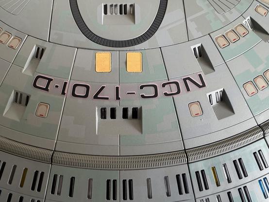 Close-up of a model of a starship, featuring the designation "NCC-1701-D" and displaying various details including windows and colored panels. The surface has a textured finish with a mix of green and gray hues.