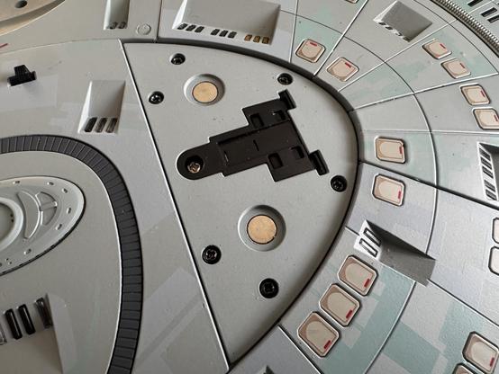 Close-up view of a detailed model or replica of a futuristic spacecraft, featuring various buttons, panels, and a central connection point. The surface has a light gray color with red and black accents.