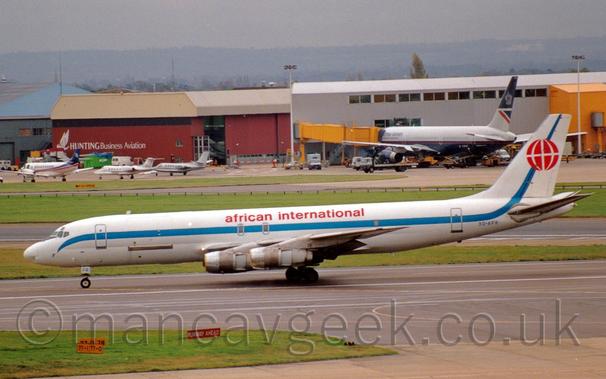Side view of a 4 engined jet airliner that has been converted to a freighter, taxiing from right to left along a wide, almost black taxiway.
The plane is mostly white, with a blue stripe running along the body and sweeping up into the tail, and red, lower-case "African International" titles on the upper fuselage above the wings.
The blue registration 3D-AFR is on the lower rear fuselage, under the tail, which itself has a stylised red globe overlaid on the diagonal blue stripe.
An strip of green grass runs halfway across the frame from the left in the foreground, while a grey terminal building with a dark blue and grey jet airliner hooked up to a yellow airbridge comes halfway across the background from the right, and a red hangar with a sandy brown roof and a brace of BizJets and a bizprop in front on the left.
Grey sky fills the rest of the image.