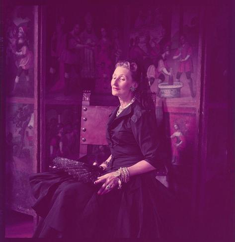 This image is a portrait photograph of Señora Julia Frazer Luckie de Diez Hidalgo, taken in the late 1950s. The woman is dressed elegantly in a dark gown and adorned with jewelry including earrings, bracelets, rings, and a necklace. She has styled her hair up with curls framing her face.

The background features colorful murals or paintings depicting various scenes, likely historical or cultural events. The overall color tone of the image appears to be purple-hued due to the lighting or post-processing effects used by the photographer. 

This photograph was taken during a time when Toni Frissell worked for the U.S. Department of State (1951-26), suggesting that it might have been captured as part of documenting official visits or events in Peru.

Señora Julia Frazer Luckie de Diez Hidalgo was an influential Peruvian socialite and philanthropist, known for her connections and charitable activities during the mid-20th century. The exact dates of her life are not provided; however, she is believed to have been active around that period.

The photograph depicts Señora Julia Frazer Luckie de Diez Hidalgo in a formal setting, possibly within an important building or establishment showcasing Peruvian art and culture, further emphasizing her status as a prominent figure in the society of Lima, Peru.