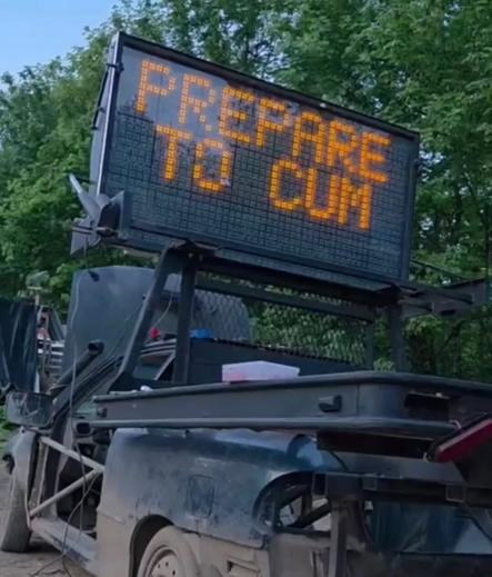 A roadside electronic construction sign displays the phrase “PREPARE TO CUM” in bright orange letters, clearly glitched or tampered with. The sign sits on a dusty trailer rig against a backdrop of trees, giving the whole scene the energy of accidental chaos.
