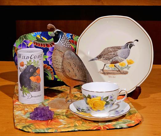 Tea tableau with a theme of California Quail, depicted on a decorative plate and as a metal sculpture, with a colorful abstract tea cozy, vintage floral china, and a chunk of amethyst geode, upon a quilted mat with an Autumn leaves pattern. The tea is a cannister of Wild Coast herbal tea with an image of a California brown bear and California poppies