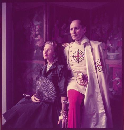In this color photograph by Toni Frissell, a man and woman are seated side-by-side in front of painted portraits. The woman is wearing an ornate black dress with pearl jewelry, holding a fan made of feathers or sequins. She has her hair up and wears makeup including eyeliner and lipstick.

The man on the right is dressed as if he's partaking in some sort of ceremonial event, sporting white pants and boots along with what appears to be armor plates over his chest that are adorned with a cross design featuring red lines intersecting at various angles. His cape has gold buttons running down its length, suggesting it might have been made from velvet or another rich fabric.

The man's jacket is embellished further by the addition of decorative shoulder straps and he holds something in both hands which could possibly be gloves and a ceremonial item such as an orb or scepter but not entirely clear. His expression seems serious with his mouth slightly open, perhaps mid-conversation.

In terms of colors, there are muted purples throughout the photo likely due to either artificial lighting at that time period (the late 1940s/early '50s) or an intentional artistic effect by Frissell herself.