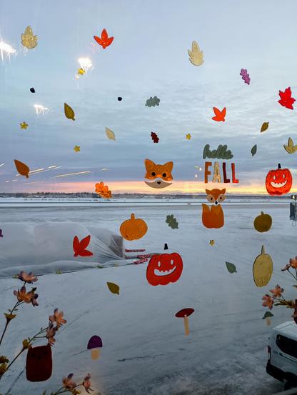 A scene outside an airport window of a flat white landscape with frosted trees in the background and an early winter sunset beneath the clouds in progress. The window is decorated with sticky gel leaves, jack-o-lanterns, foxes, and mushrooms.