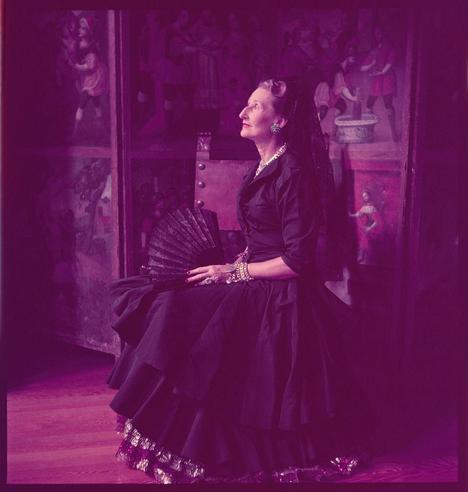 A woman wearing a long black dress and carrying an ornate fan sits in front of paintings on the wall. The photograph has a purple tint, which adds to its vintage look. The image is part of the Toni Frissell Photograph Collection donated to the Library of Congress in 1984. This collection features photographs by American photographer Toni Frissell and includes portraits of individuals such as Señora Julia Frazer Luckie de Diez Hidalgo, a Peruvian socialite and philanthropist who was born in Lima, Peru, and married Sir Henry Hurd Luckie de Diez Hidalgo.