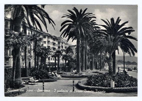 The image is a black and white photograph depicting an urban scene with palm trees, benches, and people relaxing. The setting appears to be a public garden or promenade adjacent to buildings that resemble residential apartments or hotel accommodations. There are several tall palm trees distributed throughout the area, providing shade along pathways lined with decorative bushes. Benches offer seating for visitors, some of which are occupied by individuals who seem to be enjoying their surroundings.

In the background, multi-story apartment-style buildings can be seen, suggesting an urban environment possibly in a coastal city due to the presence of palm trees and promenade-like walkways. The sky is overcast with clouds, contributing to a somewhat muted lighting effect across the scene. There's visible text overlaid on the image which reads "Riviera dei Fiori - Sanremo - I palmiri di Corso Trento Trieste," indicating that this location may be associated with or named after these places.

The overall atmosphere conveyed by the photograph is one of leisure and tranquility, typical for a promenade area in an Italian Riviera town known for its scenic beauty. The absence of vibrant colors due to the black-and-white format adds a timeless quality to the scene.
