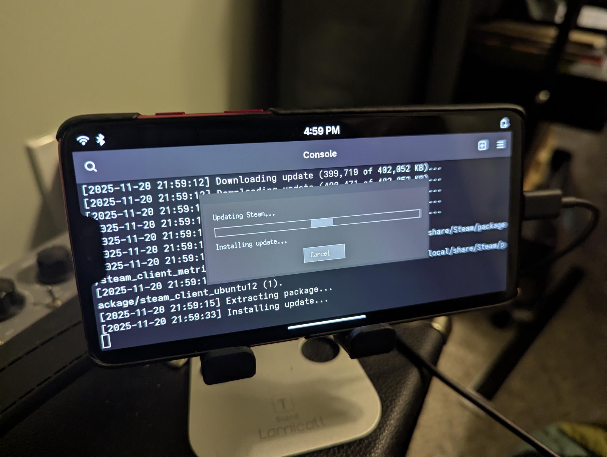 Photo of a phone on a stand running PostmarketOS, displaying a terminal with a logs and Steam's grey "Updating Steam" window.
