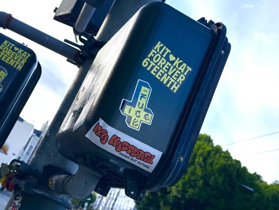 Here is alt-text for the image you provided:

Alt-text:
A close-up of a dark green pedestrian-crosswalk signal control box on a street corner, covered with stickers. One yellow sticker reads “KIT ♥ KAT FOREVER 6TEENTH” in bold block letters. Another sticker, shaped like a Dog Town cross, has stylized text “FUCK” and “ICE” in a scribbled, graffiti-like style. Below it is a white sticker with bubbly orange lettering that reads “PIG HEROSSS” followed by the smaller tagline “ONLINE BUT OUTSIDE.” The scene is slightly angled upward, showing part of the metal pole, another stickered control box to the left, and green trees and a bright blue sky in the background.