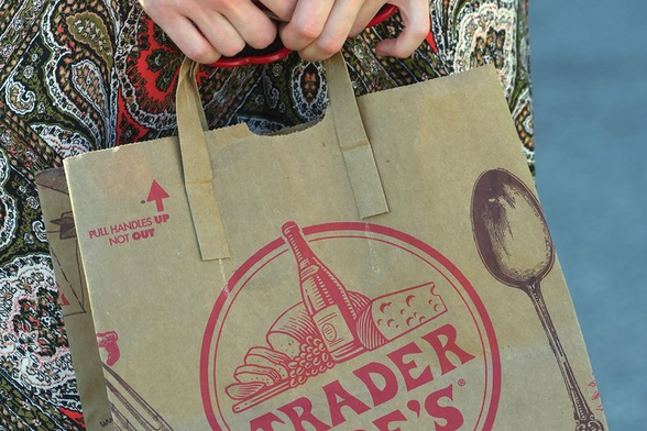 Close-up of a person holding a Trader Joe’s brown paper shopping bag with red logo and illustrations of a bottle, cheese, and bread. The person’s hands grip the bag handles, and they are wearing a patterned shirt in the background.