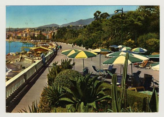 The image depicts a scenic view of the Riviera dei Fiori, with lush greenery and tropical plants prominently featured in the foreground. The middle ground shows an outdoor seating area under large yellow and blue striped umbrellas, surrounded by empty lounge chairs that suggest a leisurely atmosphere at what appears to be a beachside promenade or restaurant terrace.

In the background, there is a bustling seaside town with colorful buildings alongside shops and eateries lining the waterfront. People are seen walking along the boardwalk and engaging in various activities such as sitting on benches or standing by the railings overlooking the water. The setting sun casts warm hues over the architecture of the town, emphasizing its vibrant atmosphere.

The landscape includes a mix of urban elements like street lamps and modern infrastructure juxtaposed with natural scenery consisting of palm trees and mountainous terrain extending into the distance under a clear blue sky. This blend creates an inviting tableau that captures both the charm of coastal life and the allure of Mediterranean living.