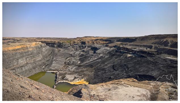 A panoramic, wide angle photograph in colour, signed by Kev Peirce, showing a massive open cut mine.  The image captures the immense scale of the excavation, with distinct, stepped layers (benches) visible along the sides of the pit, highlighting the geology and mining method.  The base of the mine is a mix of dark, excavated material and two pools of stagnant, yellowish green water. The surrounding landscape is arid and flat under a clear, pale blue sky, emphasising the isolation and the environmental impact of the operation.