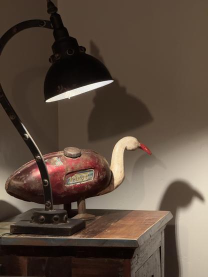 A vintage lamp casts light on a whimsical decorative piece resembling a duck, with a body made of an old metal motorcycle gas tank with a red and white color scheme, set on a wooden surface. The lamp and the duck cast shadows on the wall behind.