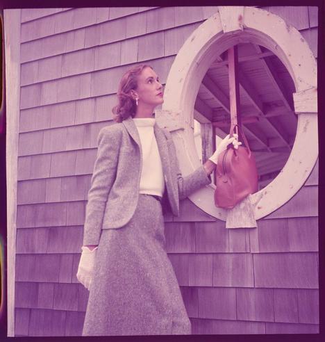 a woman standing beside a round porthole window on the side of a house and looking up as she is about to put her red handbag through it. The photograph has been printed in sepia tones, which give it an old-fashioned look. It could be considered one of Toni Frissell's works from around 1951-1952 based on the fashion styles shown here.