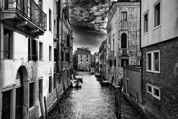 Schwarz Weiss Aufnahme eines Kanals in Venedig. Links und rechts erkennt man die Rückseite von Häusern, manche haben kleine Balkone. Im Hintergrund ist ein weiteres Haus zu erkennen und ein Stückchen des Himmels mit weissen Wolken.