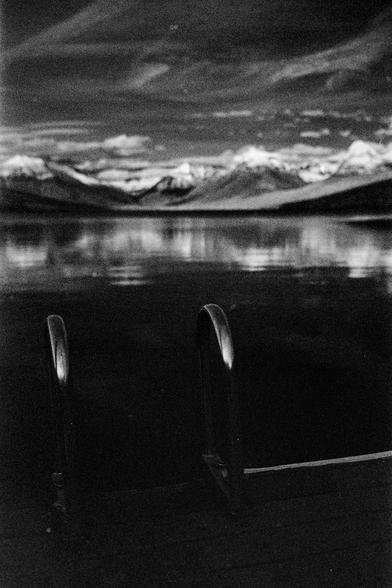A black and white photo with a pair of raised ladder handles attached to the end of a dock which is sitting on a lake that stretches to the out of focus background framed by mountains with snowy peaks under a darker sky with swirly white clouds.