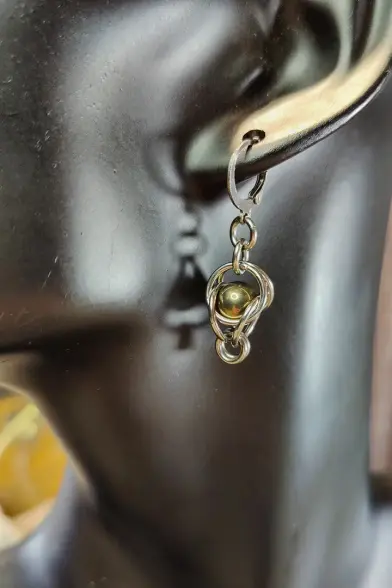 Boucle d'oreille montrant une perle qui semble magiquement tenir entre deux paires d'anneaux, comme posée à la jonction. La perle est de couleur bronze.