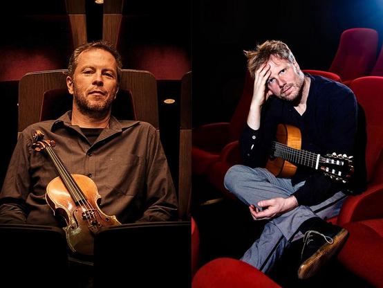 Zwei Männer sitzen auf roten Stühlen, einer hält eine Geige, der andere eine Gitarre. Im Hintergrund ein dunkler Raum.