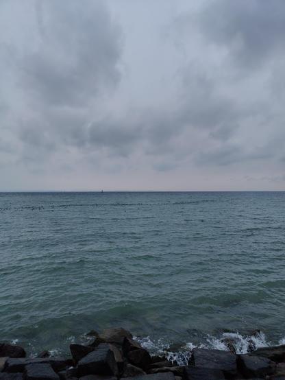 Blick von Romanshorn Hafen Richtung Osten über den Bodensee, dichte Wolkendecke, das andere Ufer fast nicht zu sehen, türkisfarbenes Wasser mit leichten Wellen, die Leuchtturmdalbe links der Bildmitte, ein Schwarm Wasservögel auf dem Wasser, im Vordergrund Steinblöcke der Uferbefestigung im Wasser
