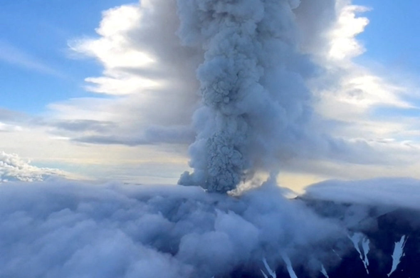 Éruption du volcan Krasheninnikova, avec un panache de cendres et de gaz s'élevant dans le ciel bleu, entouré de nuages.