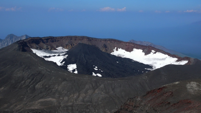 Vue aérienne du volcan Krasheninnikova, montrant son cratère plein de neige et de cendres noires, avec des sommets montagneux en arrière-plan.