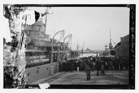 The image depicts a historical scene, likely from the early 20th century based on the black and white film quality. A large naval ship is docked in a port with its hull visible displaying multiple gun turrets and what appears to be artillery pieces along its sides. Several cranes are seen alongside the ship, indicating an active industrial or military environment.

A significant crowd of people has gathered around the ship's perimeter, suggesting it may have been of great interest at that time, possibly related to a royal event as mentioned in the caption regarding King Faisal II of Iraq arriving in Haifa. Some individuals appear dressed in naval uniforms while others are civilians wearing hats and coats typical of early 20th-century fashion.

The image quality is somewhat deteriorated with visible scratches or damage on its surface, which adds to the historical ambiance but obscures some finer details within the photograph itself. The overall impression conveys a moment of public significance at an industrial port during a bygone era.