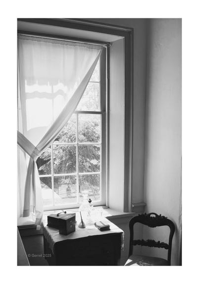 Black and white photo of a vintage room. Sunlight through a window casts shadows on a table holding a wooden box and glass bottle. A chair sits nearby.