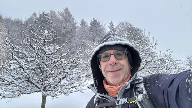 Selfie of me, a middle-aged white man with glasses smiling with snow on my hood and shoulders against a backdrop of snowy pasture and snow-covered trees.