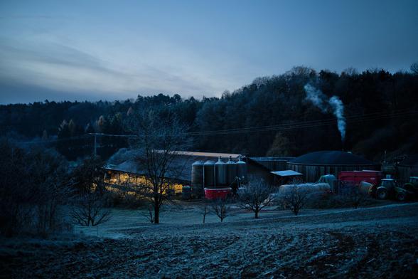 Ein beleuchteter Stall in der Ferne. Große Behälter aus Metall. Am Anfang. Rechts die Biogas Behätnisse mit gebogenen Dach aus Gummi. Ballen Trecker und Ahnhänger rechts an der Seite. In Vordergrund ein Feld. Im Hintergrund Wald. Eine Strohmleitung übern Stall. Rauch vom Motor bei den Gas Behältern. Im Bild alle smit Frost bedeckt.