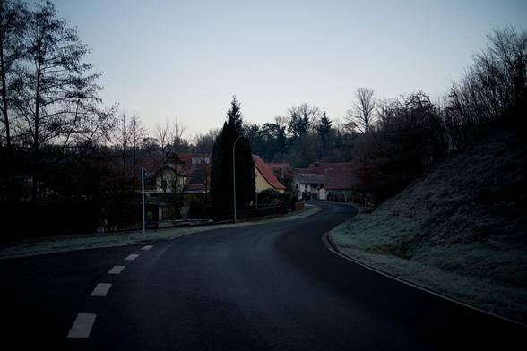 Die Straße Schlängelt sich durchs Bild. Rechts ein Ranga. (Hang) Mit Gestrüpp. Der in Häuser Übergeht in der Ferne. Links Gärten. Danach auch Häuser. Im Vordergrund geht eine Straße links ab. Überall vereinzelt Bäume.