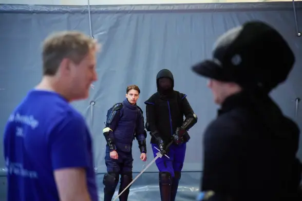 Two longsword-armed HEMA fencers in protective gear prepare for a bout as others watch.