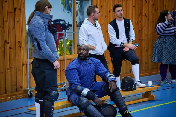 A group of HEMA participants relax and chat on a bench, one wearing full blue protective gear.