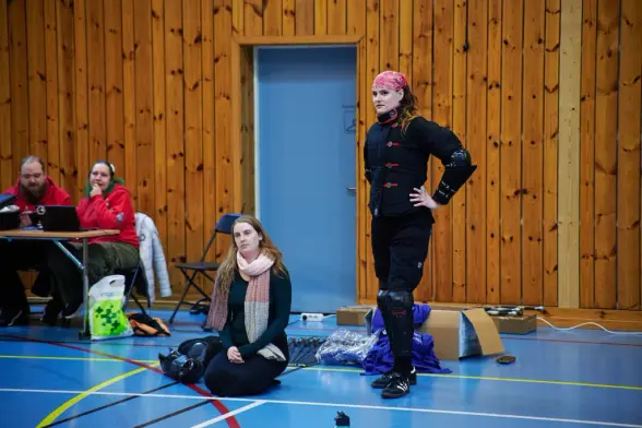A seated spectator watches while a HEMA fighter in black gear stands ready beside the judges’ table.