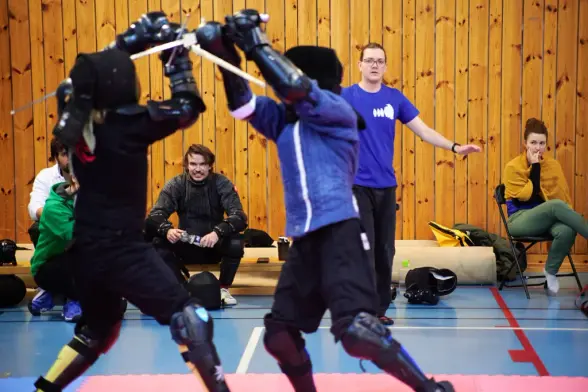 Two HEMA fighters exchange overhead strikes as a referee watches the action closely.